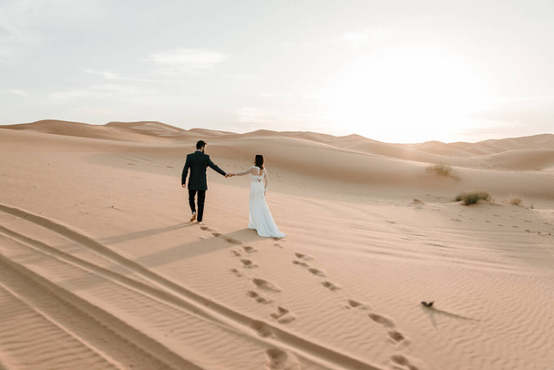 Ein Paar im Brautkleid läuft im Morgengrauen barfuß durch die Wüste. Hochzeitskonzept - Foto, Bild