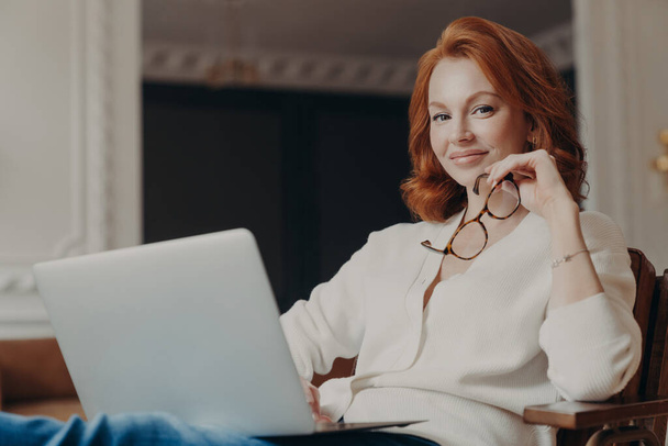 Young beautiful redhead skilled woman sends file to customer via modern laptop, sits in comfortable armchair, holds eyewear, spends time on online education, watches webinars, has pleased expession - Photo, Image