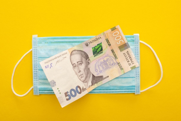 Sarı arka planda Ukrayna banknotu ve tıbbi maskesinin üst görüntüsü - Fotoğraf, Görsel