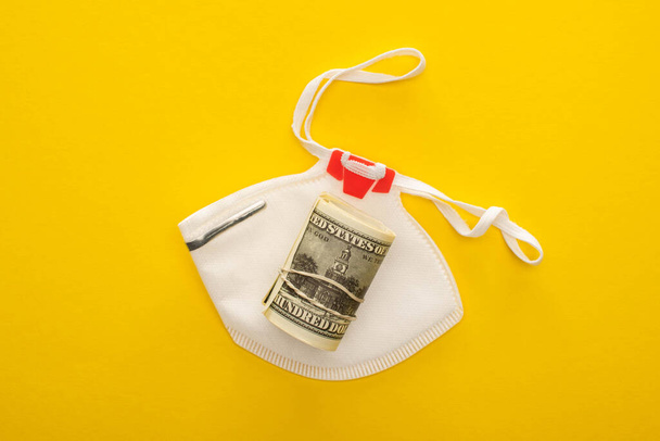 κορυφαία άποψη του roll χρήματα και μάσκα ασφαλείας σε κίτρινο φόντο - Φωτογραφία, εικόνα