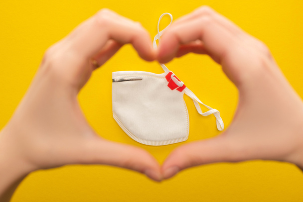 cropped view of woman showing heart with hands above safety mask on yellow background - Foto, Bild