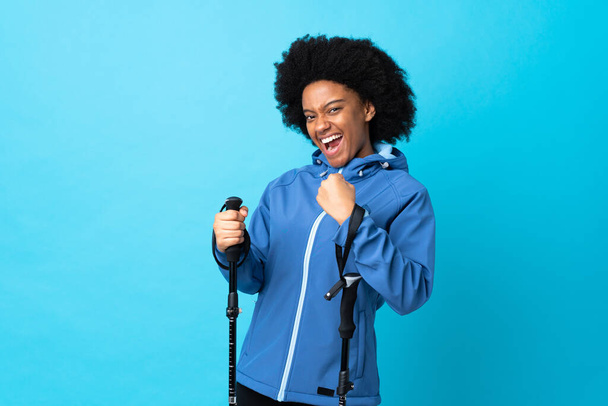 Young Africa American with backpack and trekking poles isolated on blue background celebrating a victory - 写真・画像