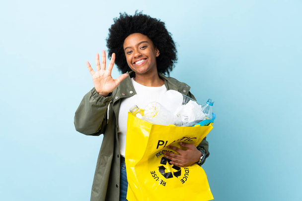 Молода афро - американська жінка, яка тримає пакет з переробкою, ізольований на кольоровому фоні, рахуючи п "ять пальцями. - Фото, зображення