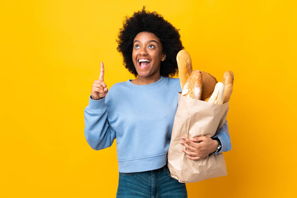 Jonge Afro-Amerikaanse vrouw koopt iets brood geïsoleerd op gele achtergrond wijzen en verrast - Foto, afbeelding