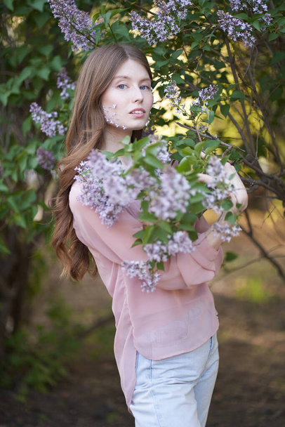 Una giovane ragazza con lunghi capelli ondulati e occhi azzurri, in piedi nel fogliame di alberi di lilla sulle guance della ragazza petali lilla, guarda direttamente nella fotocamera
                                      - Foto, immagini
