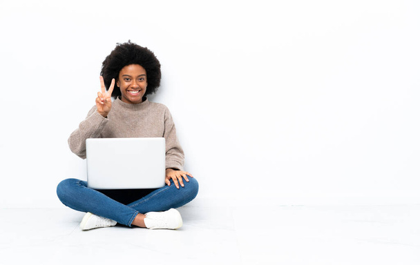 Nuori afroamerikkalainen nainen kannettavan tietokoneen kanssa istuu lattialla hymyillen ja osoittaen voitonmerkkiä - Valokuva, kuva