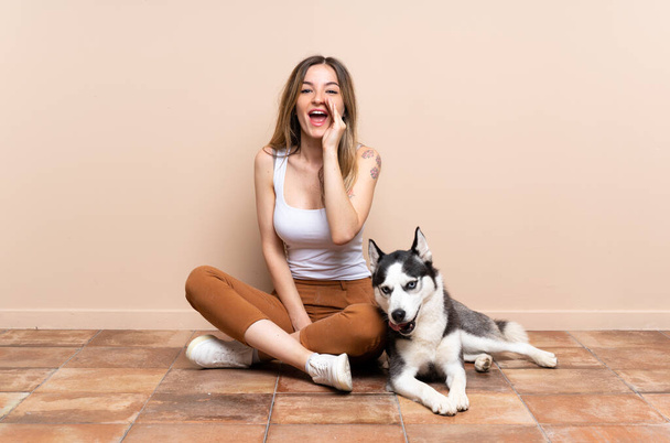 Young pretty woman with her husky dog sitting in the floor at indoors shouting with mouth wide open - 写真・画像