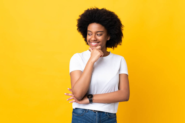 Молодая афроамериканка изолирована на желтом фоне, смотрит в сторону и улыбается - Фото, изображение