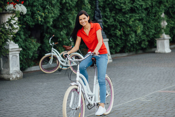 Menina sorridente bonita em uma camiseta vermelha monta uma bicicleta branca
 - Foto, Imagem