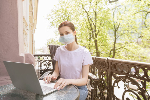 young woman in medical mask sitting on chair and using laptop on balcony  - Фото, зображення