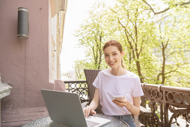 gelukkig en aantrekkelijk freelancer houden smartphone en het gebruik van laptop op het balkon  - Foto, afbeelding