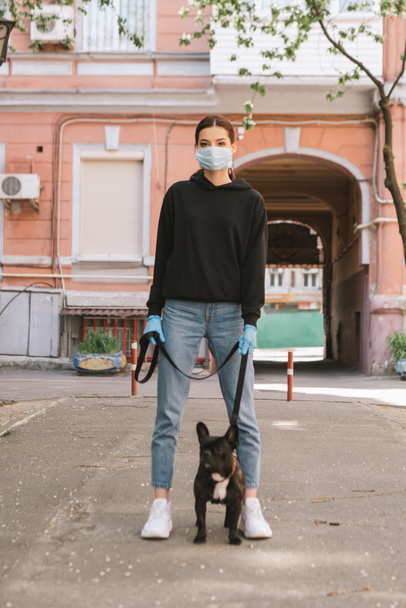 giovane donna in maschera medica e guanti di lattice in piedi con carino bulldog francese sulla strada
  - Foto, immagini