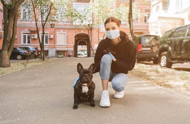 Nainen lääketieteellinen naamio ja lateksi käsineet koskettaa söpö ranskalainen bulldog kadulla  - Valokuva, kuva