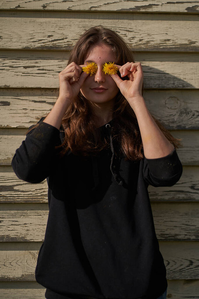  Uma jovem segurando flores amarelas na frente de seus olhos. Contra o fundo de uma casa de madeira
                                 - Foto, Imagem