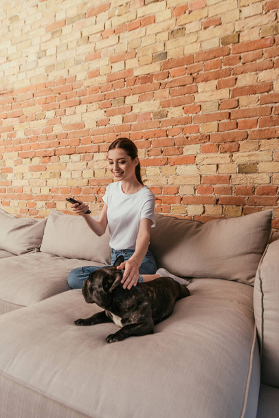 heureux jeune femme tenant télécommande et assis sur canapé avec bulldog français
 - Photo, image
