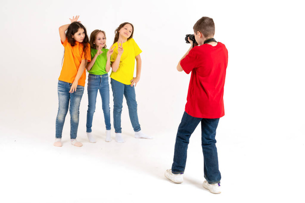 A boy photographer takes pictures of three cute girls in bright T-shirts on a white background in the studio. Young photographer and blogger - Photo, Image