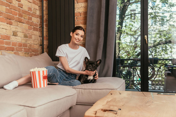 selective focus of cheerful girl sitting on sofa near popcorn bucket and touching cute french bulldog  - Фото, изображение