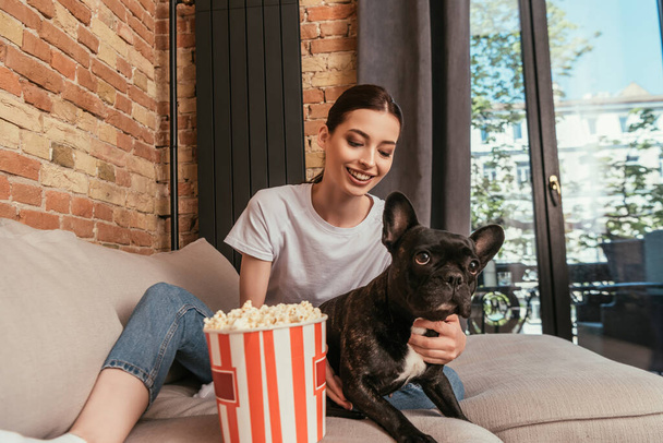 fröhliches Mädchen sitzt auf Sofa neben Popcorn-Eimer und berührt niedliche französische Bulldogge  - Foto, Bild