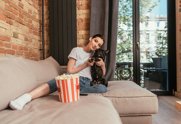 fröhliches Mädchen sitzt auf Sofa neben Popcorn-Eimer und umarmt niedliche französische Bulldogge  - Foto, Bild