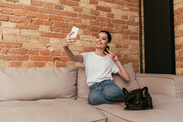 gelukkig vrouw tonen vrede teken terwijl het nemen van selfie met leuke franse bulldog in de woonkamer  - Foto, afbeelding