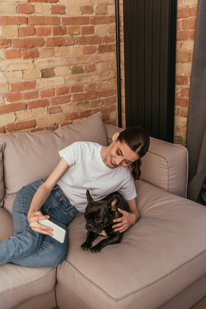 cheerful young woman taking selfie with cute french bulldog in living room  - Foto, immagini