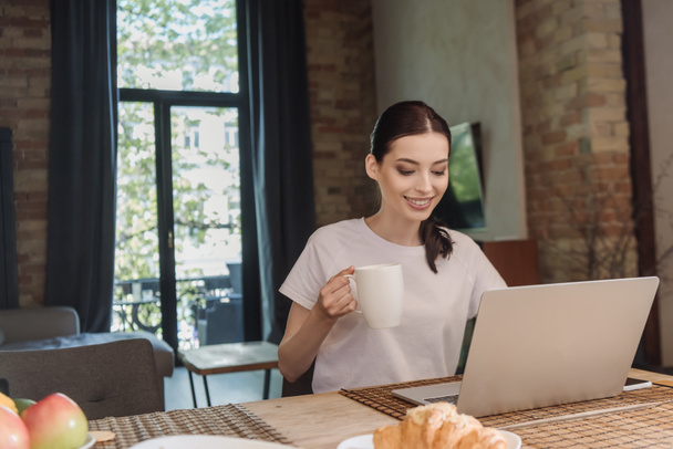 happy freelancer holding cup of coffee near laptop and croissant on table  - Photo, Image