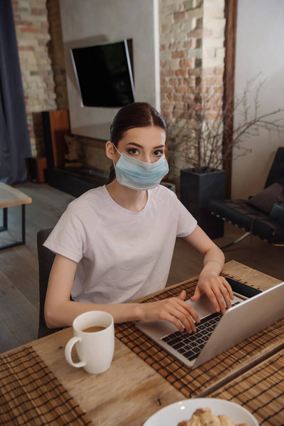 young freelancer in medical mask typing on laptop near cup of coffee - Fotó, kép