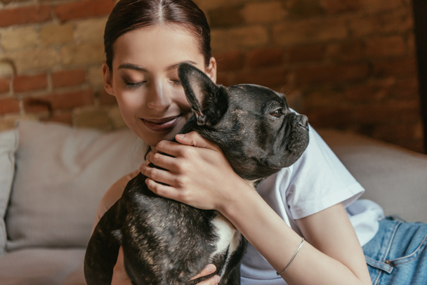 young woman with closed eyes touching black french bulldog at home - Photo, image