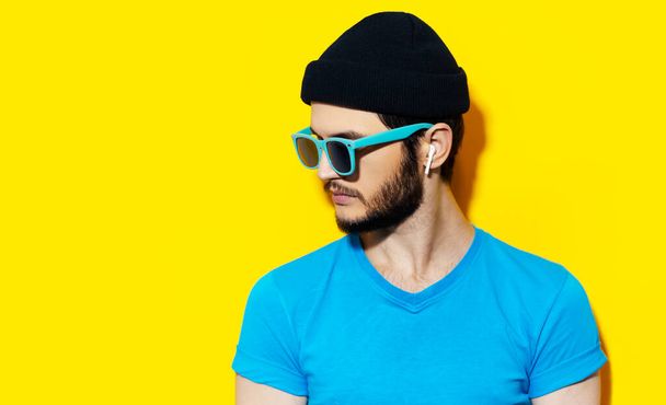 Studio portrait of young hipster guy using wireless earphones, wearing blue shirt, cyan sunglasses and black beanie hat on background of yellow color with copy space. - Photo, Image