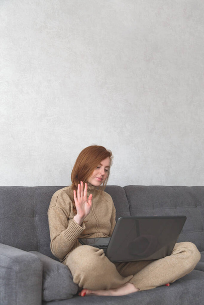 Jonge roodharige vrouw in casual kleding is met behulp van een laptop, zwaaien en glimlachen terwijl video chatten. - Foto, afbeelding