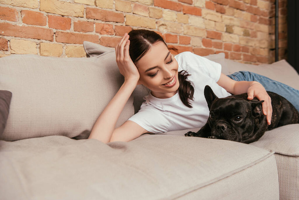 onnellinen tyttö makaa sohvalla ja koskettaa musta ranskalainen bulldog  - Valokuva, kuva