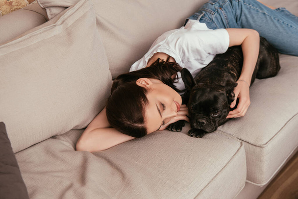 веселая девушка лежит на диване и трогает черного французского бульдога
  - Фото, изображение