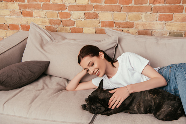 Gülümseyen kız kanepede uzanıyor ve siyah Fransız bulldog 'una dokunuyor.  - Fotoğraf, Görsel