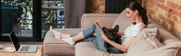 panoramic crop of happy young woman reading book near black french bulldog and laptop with blank screen  - Fotografie, Obrázek