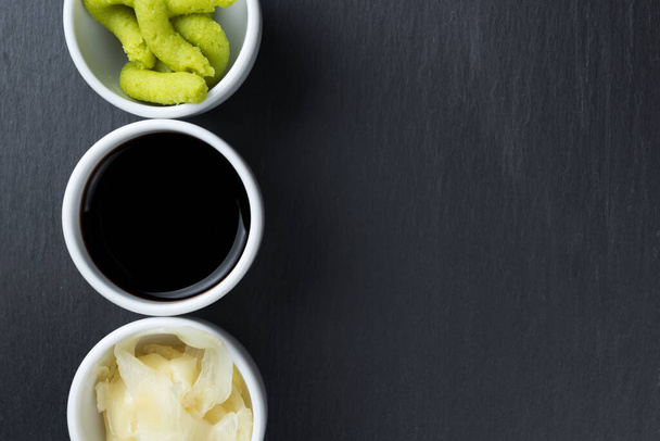 Japanischer Meerrettich-Wasabi, Sojasauce und Ingwer in kleinen weißen Schalen auf einem schwarzen Küchenteller. Kopierraum auf der rechten Seite. Abstraktes Konzept der asiatischen Küche. - Foto, Bild