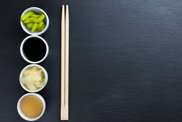 Soja en vissaus, gember, Japanse mierikswortel wasabi in kleine witte kommen met eetstokjes op zwarte keukenblad achtergrond. Typische Aziatische keuken kruiden. Kopieer ruimte aan de rechterkant. - Foto, afbeelding