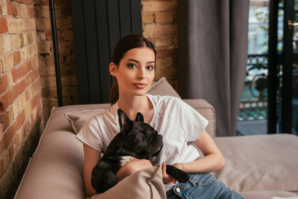 attractive young woman holding cute french bulldog and looking at camera - Photo, Image