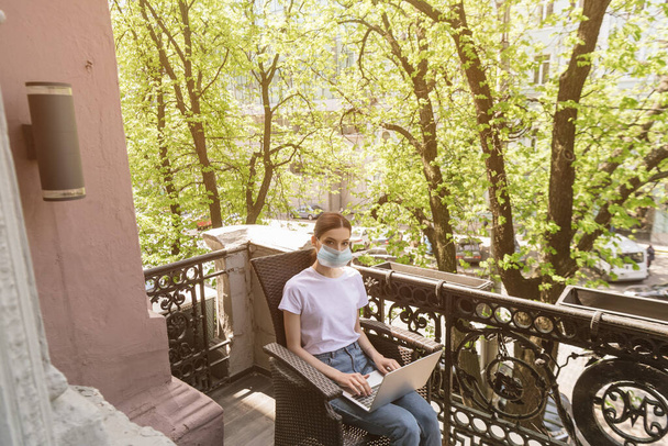joven freelancer en máscara médica sentado en la silla, utilizando el ordenador portátil y mirando a la cámara en el balcón
  - Foto, Imagen