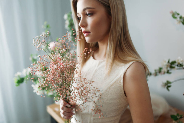 A beautiful girl with blonde hair in a wedding dress holds a branch of dyed gypsophila in her hands. Photo session of the bride in the Studio decorated with Apple trees - Photo, Image