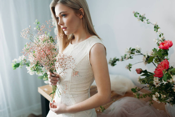 A beautiful girl with blonde hair in a wedding dress holds a branch of dyed gypsophila in her hands. Photo session of the bride in the Studio decorated with Apple trees - Fotó, kép