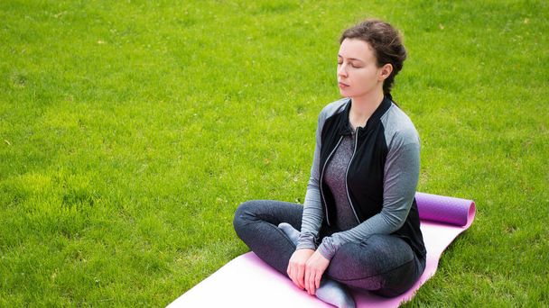 ヨガフィットネススポーツウェアを身に着けている白人女性ヨガを練習,緑の芝生の上の公園で目を閉じて屋外.マットの上にハスの位置に座って強度トレーニングワークアウトの前に穏やかな選手瞑想 - 写真・画像