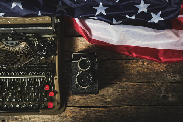 Old retro typewriter, a vintage camera, and the flag of the United States of America on a wooden table - Foto, Imagen