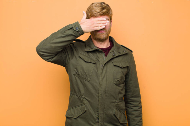 young blonde man covering eyes with one hand feeling scared or anxious, wondering or blindly waiting for a surprise against flat wall - Photo, image