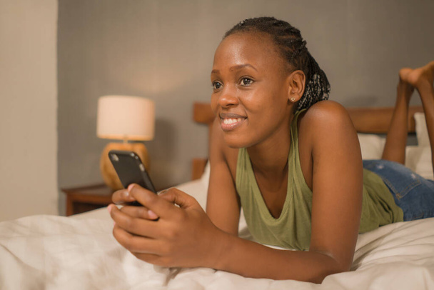 Домашній портрет молодої щасливої і привабливої чорношкірої жінки, яка лежить на ліжку, використовуючи соціальний медіа додаток в мобільному телефоні, розслаблений і веселий онлайн побачень - Фото, зображення