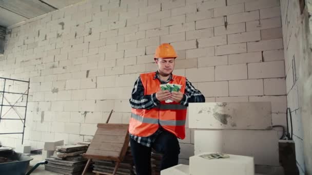 A male builder in an orange yellow helmet and an orange helmet stands indoors against a brick wall and slowly and happily counts the money. - Footage, Video