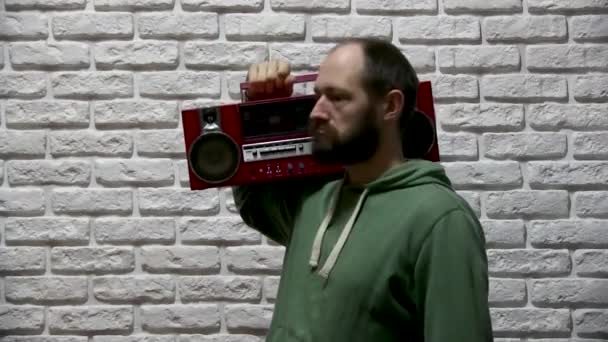 een man in een groene sweater met een capuchon en een baard staat tegen een achtergrond van witte bakstenen, pakt een rode oude grote tape recorder en zet het op zijn schouder, kijkt rond en gaat achter - Video