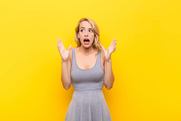 young blonde woman looking shocked and astonished, with jaw dropped in surprise when realizing something unbelievable against orange wall - Фото, изображение
