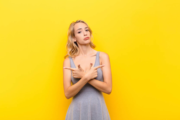 young blonde woman looking puzzled and confused, insecure and pointing in opposite directions with doubts against orange wall - Foto, imagen