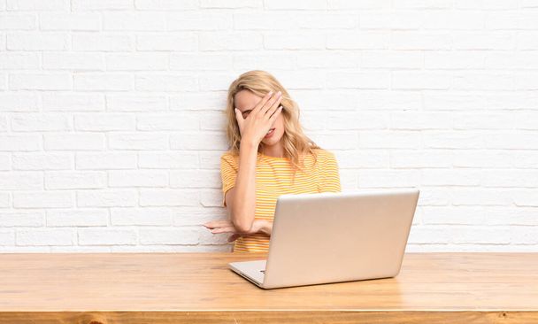 young blonde woman looking stressed, ashamed or upset, with a headache, covering face with hand using a laptop - Fotoğraf, Görsel