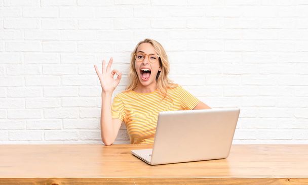 young blonde woman feeling successful and satisfied, smiling with mouth wide open, making okay sign with hand using a laptop - Zdjęcie, obraz
